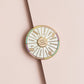 A circular enamel pin shaped like a dessert wheel, featuring gold-toned metal borders and segments labeled with sweet treats and desserts such as cake, cookies, chocolate, gummy, cupcakes, macarons, pie, and ice cream. The pin is photographed on a soft beige background with a diagonal shadow.