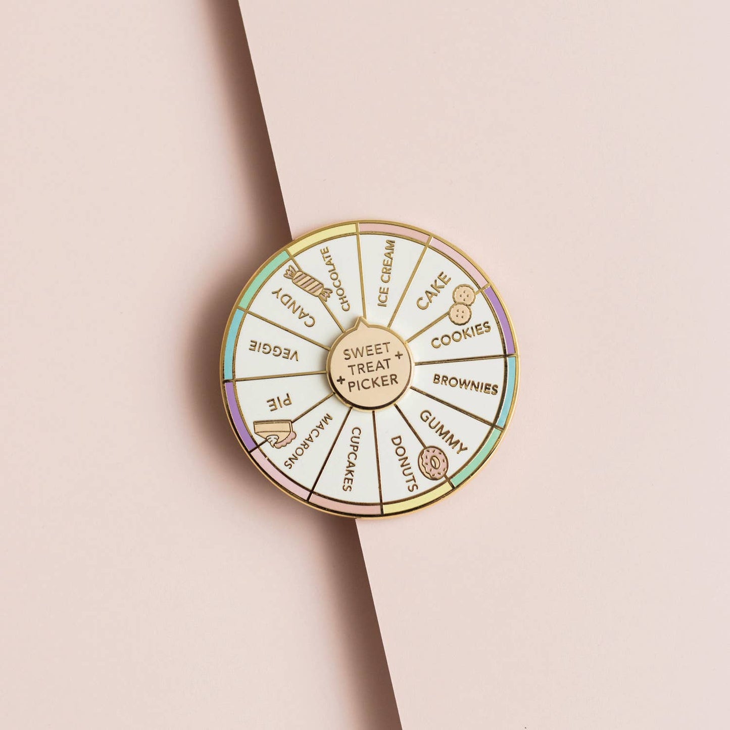 A circular enamel pin shaped like a dessert wheel, featuring gold-toned metal borders and segments labeled with sweet treats and desserts such as cake, cookies, chocolate, gummy, cupcakes, macarons, pie, and ice cream. The pin is photographed on a soft beige background with a diagonal shadow.