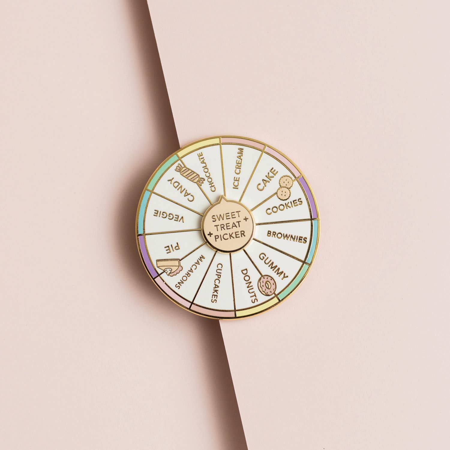 A circular enamel pin shaped like a dessert wheel, featuring gold-toned metal borders and segments labeled with sweet treats and desserts such as cake, cookies, chocolate, gummy, cupcakes, macarons, pie, and ice cream. The pin is photographed on a soft beige background with a diagonal shadow.
