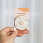 A circular enamel pin shaped like a dessert wheel, featuring gold-toned metal borders and segments labeled with sweet treats and desserts such as cake, cookies, chocolate, gummy, cupcakes, macarons, pie, and ice cream. The pin is photographed on a soft beige background with a diagonal shadow.