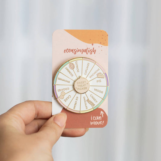 A circular enamel pin shaped like a dessert wheel, featuring gold-toned metal borders and segments labeled with sweet treats and desserts such as cake, cookies, chocolate, gummy, cupcakes, macarons, pie, and ice cream. The pin is photographed on a soft beige background with a diagonal shadow.