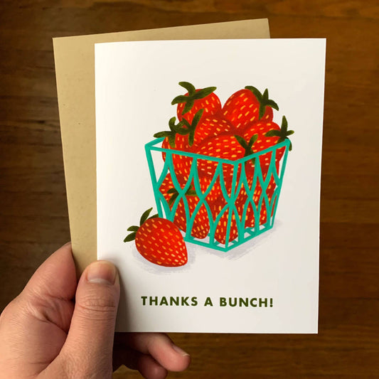 A hand holds a white greeting card featuring an illustration of red strawberries in a teal basket with the text 'THANKS A BUNCH!' below, with a kraft envelope behind it on a wooden surface.