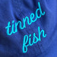 Blue hat with 'tinned fish' embroidered in turquoise on a close-up view