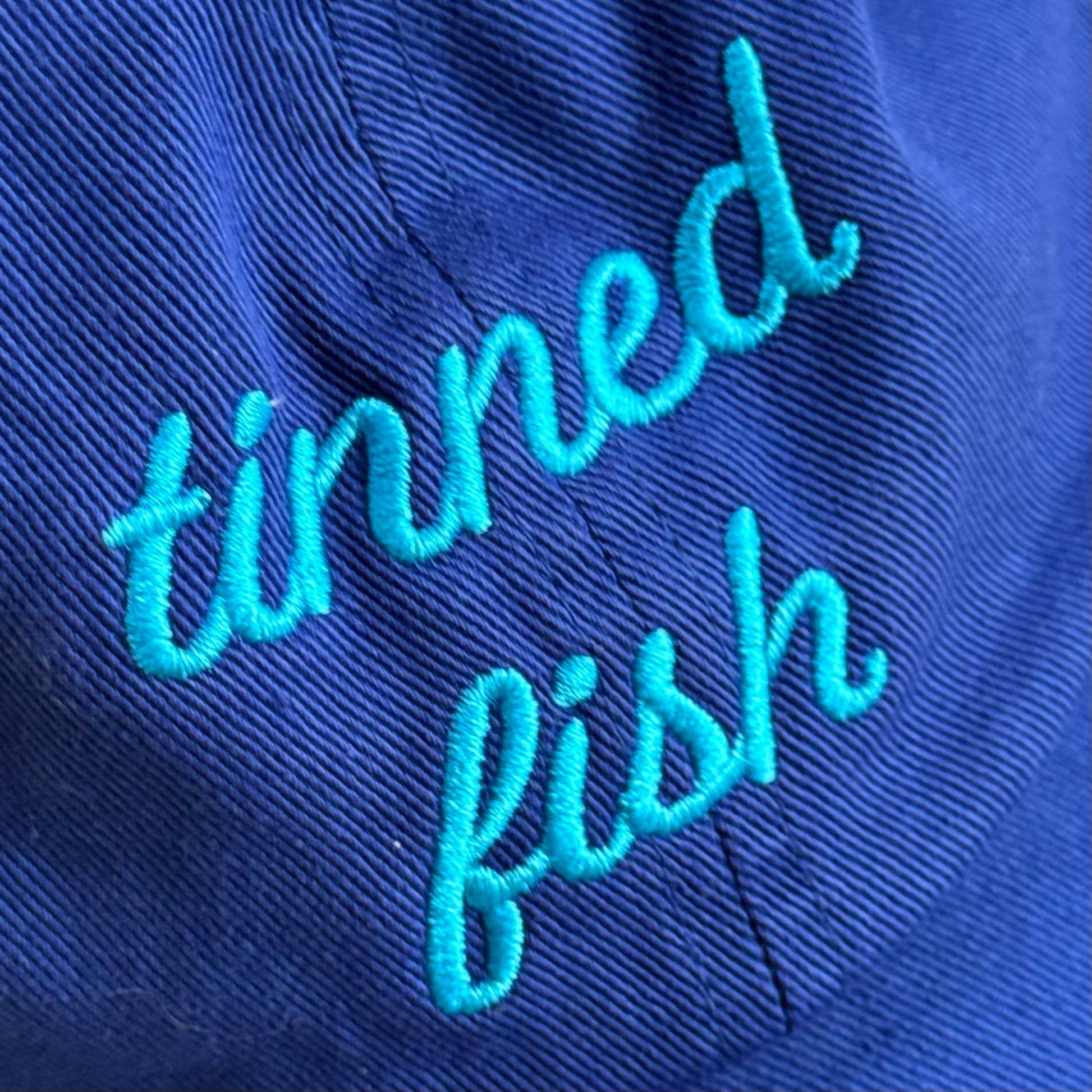 Blue hat with 'tinned fish' embroidered in turquoise on a close-up view
