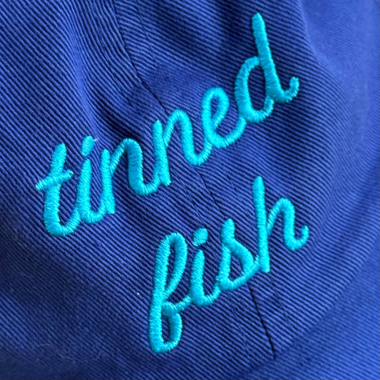 Blue hat with 'tinned fish' embroidered in turquoise on a close-up view