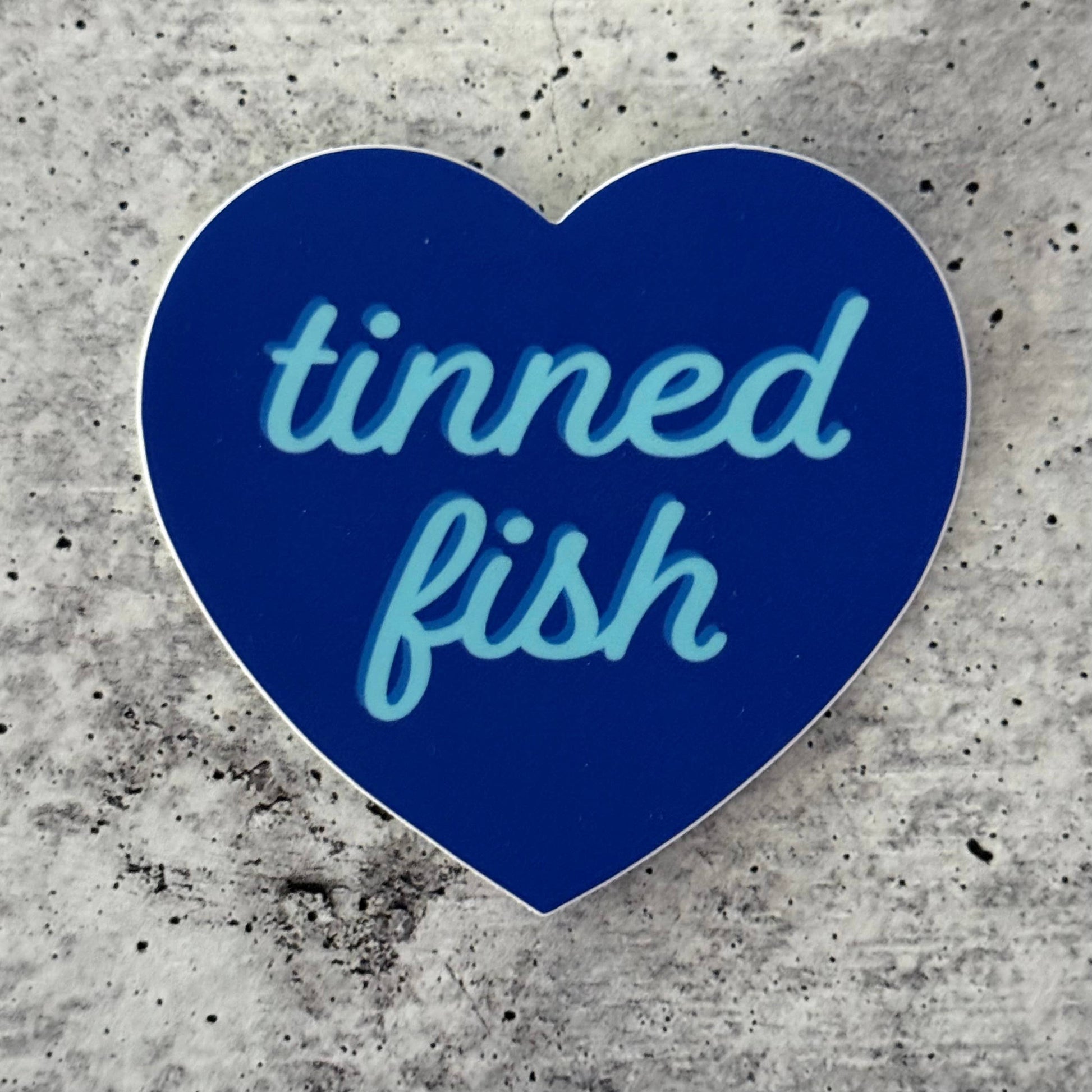 Blue heart-shaped sticker with 'tinned fish' text on a concrete surface