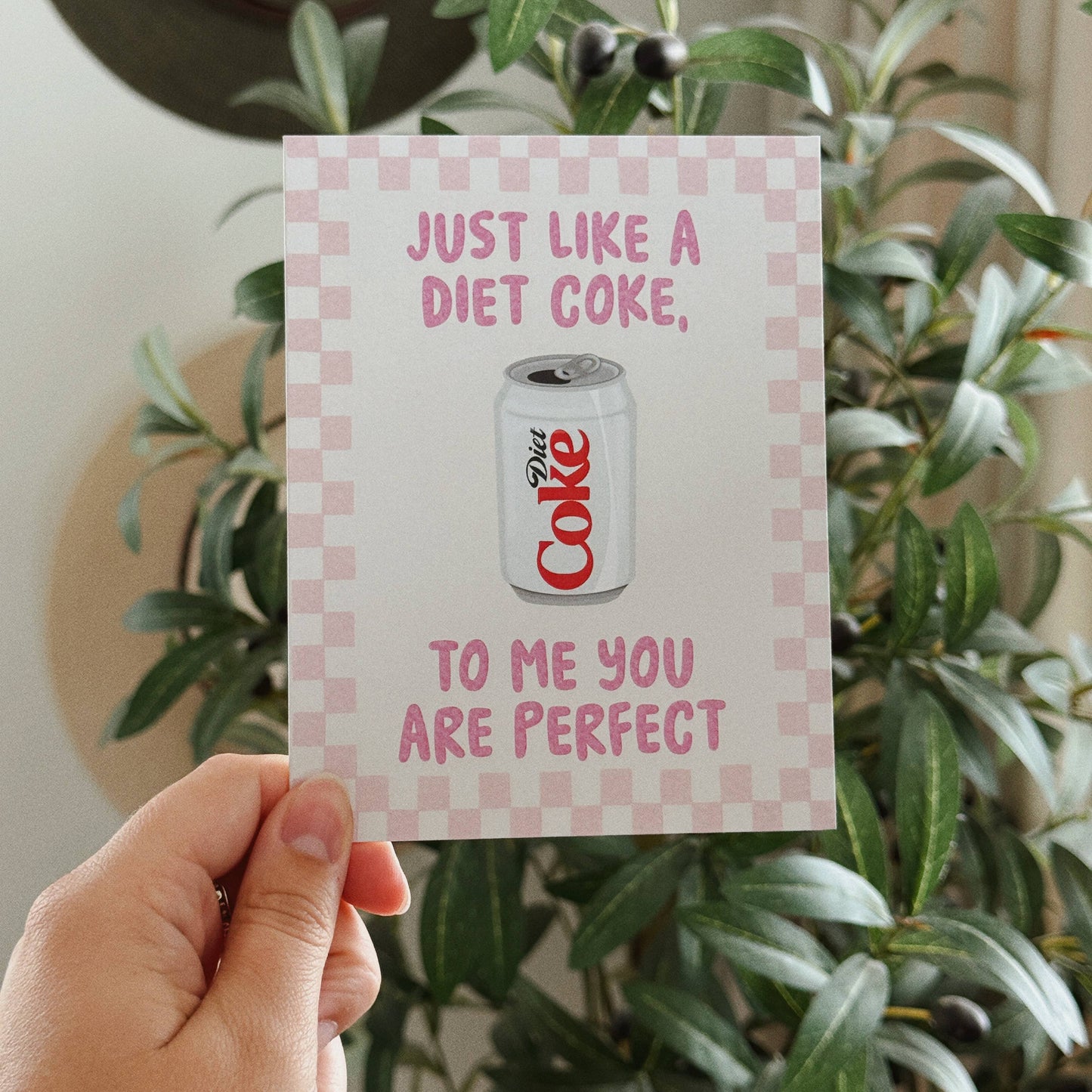 A hand holds a square greeting card in front of a green potted plant. The card has a pink and white checkered border, pink text reading 'JUST LIKE A DIET COKE, TO ME YOU ARE PERFECT', and a graphic of a Diet Coke can in the center.