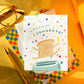 Greeting card with a toast character popping out of a toaster and 'a toast to you, Congrats!' text on a colorful background