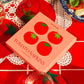 Box of tomato-themed cocktail napkins on a red tablecloth with glasses and doilies.