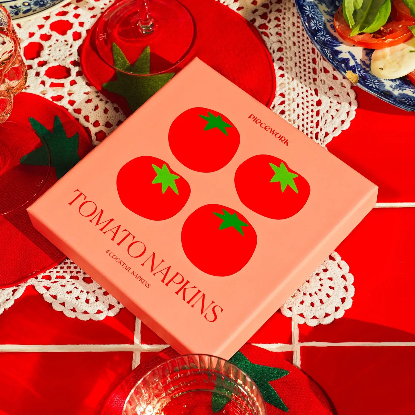 Box of tomato-themed cocktail napkins on a red tablecloth with glasses and doilies.