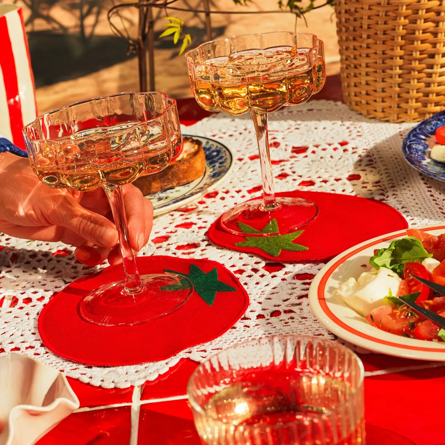 Two glasses of rose wine on red tomato shaped coasters with a table setting including a salad and a candle.