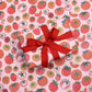 sheet of gift wrap with tomatoes all over it. On top of the sheet is a gift wrapped with the tomatoes gift wrap with a red bow 