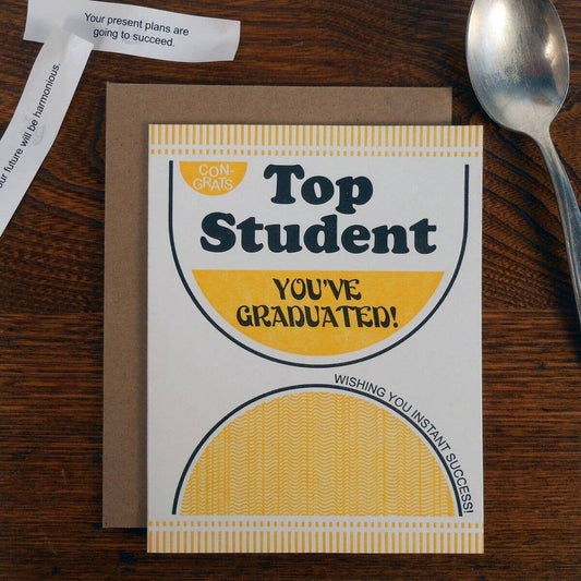 Graduation card with 'Top Student You've Graduated!' text on a wooden surface with a spoon and paper.