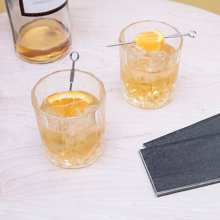 Two whiskey glasses with orange-sliced cocktails on a white tablecloth with a black napkin.
