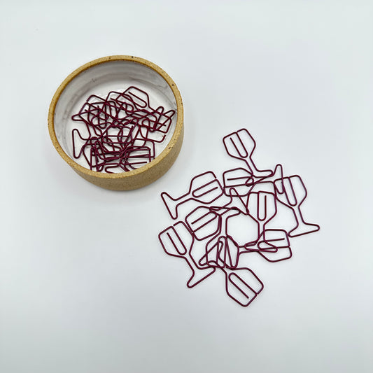 Burgundy colored wine glass paperclips in a pile on a piece of paper and in a ramekin.