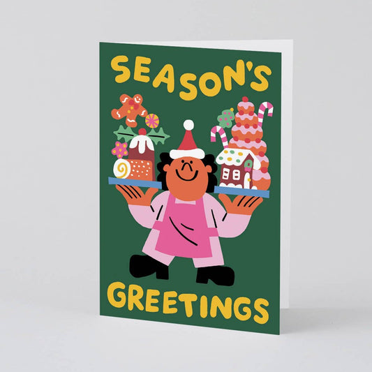 holiday greeting card that reads "season's greetings" Has illustration of a man wearing a santa hat holding two trays of holiday treats