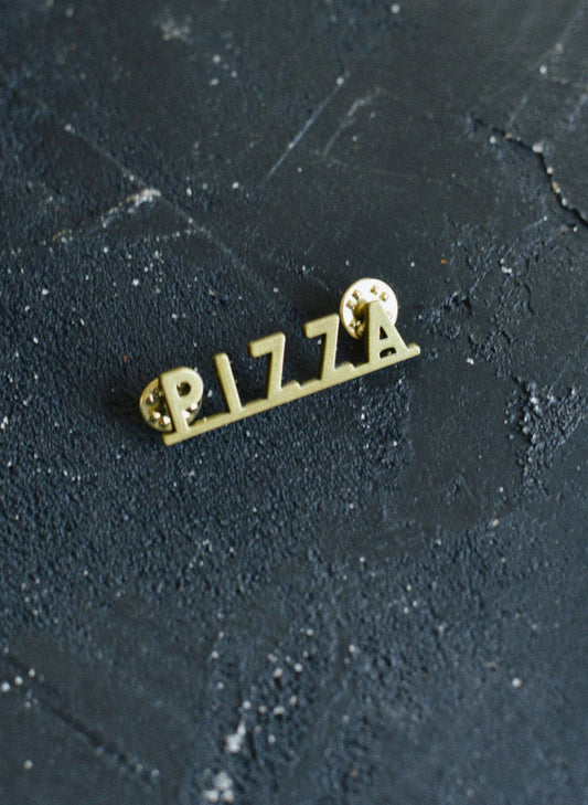 Brass colored enamel pin that spells out "pizza"