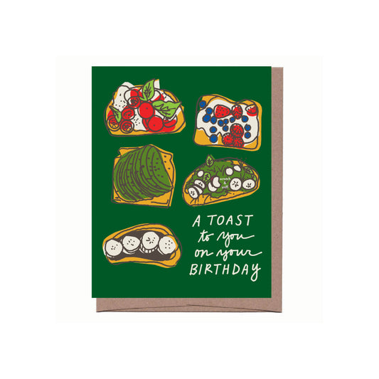 Greeting card with text "A TOAST to you on your BIRTHDAY" and illustration on vibrant green background. Illustration features five different "toasts" (ie. open-faced sandwiches) - cucumber and veggie toast, avocado toast, mixed berries and cream cheese toast, caprese toast, Nutella and bananas toast. 