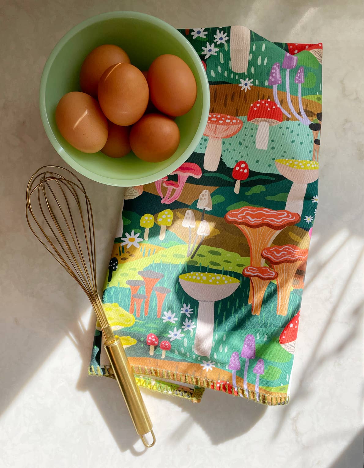 Tea towel with a colorful mushroom design all over it. Shown here with a whisk and bowl of eggs 