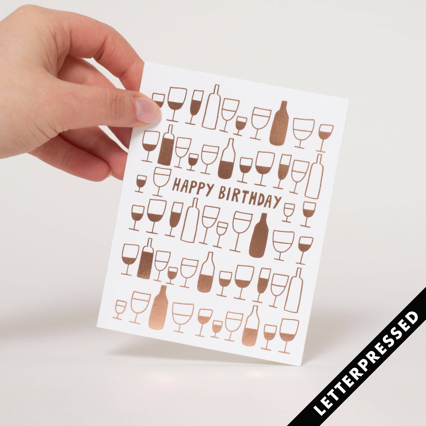 Greeting card printed in a rose gold metallic ink. Text reads "Happy Birthday" surrounded by illustration of different sized wine bottles and glasses filled at differing levels.