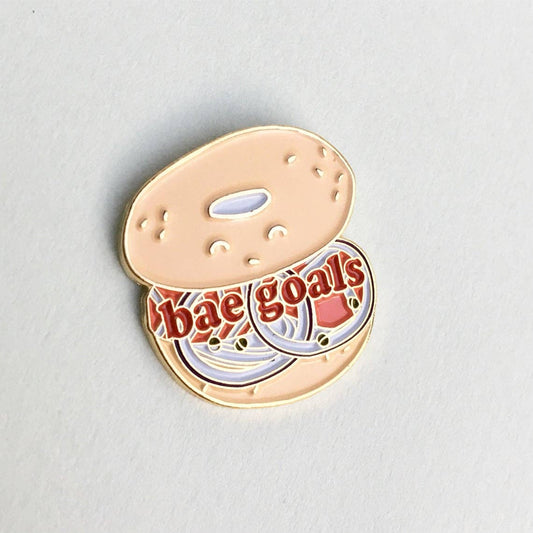 Bagel enamel pin that reads "Bae goals" on an open faced bagel. 