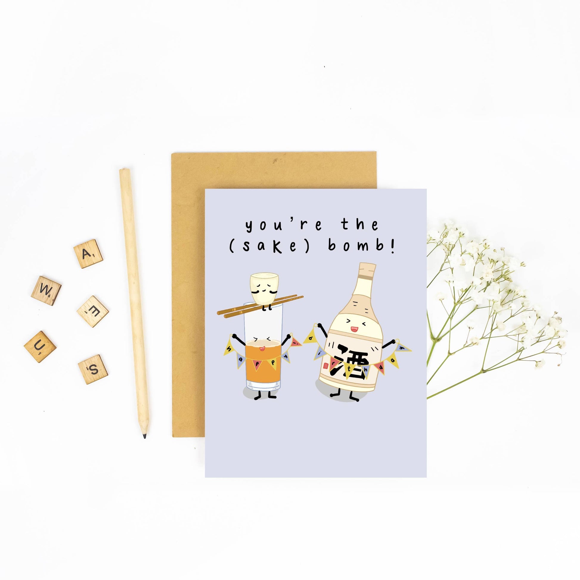 Pale blue greeting card with illustration of a happy anthropomorphic Sake Bottle holding a colorful banner that reads "happy birthday," while a Sake Cup sits precariously on top of chopsticks perched over a glass of beer. Text reads "you're the (sake) bomb!" 