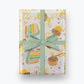 Birthday Cake Wrapping Paper shown with a blue bow. Illustration on wrapping paper features cupcakes and slices of colorful layer cake with lots of sprinkles. Multiple areas of text read "Make a WIsh!" and "Wishing you a very Happy Birthday" in gold script.