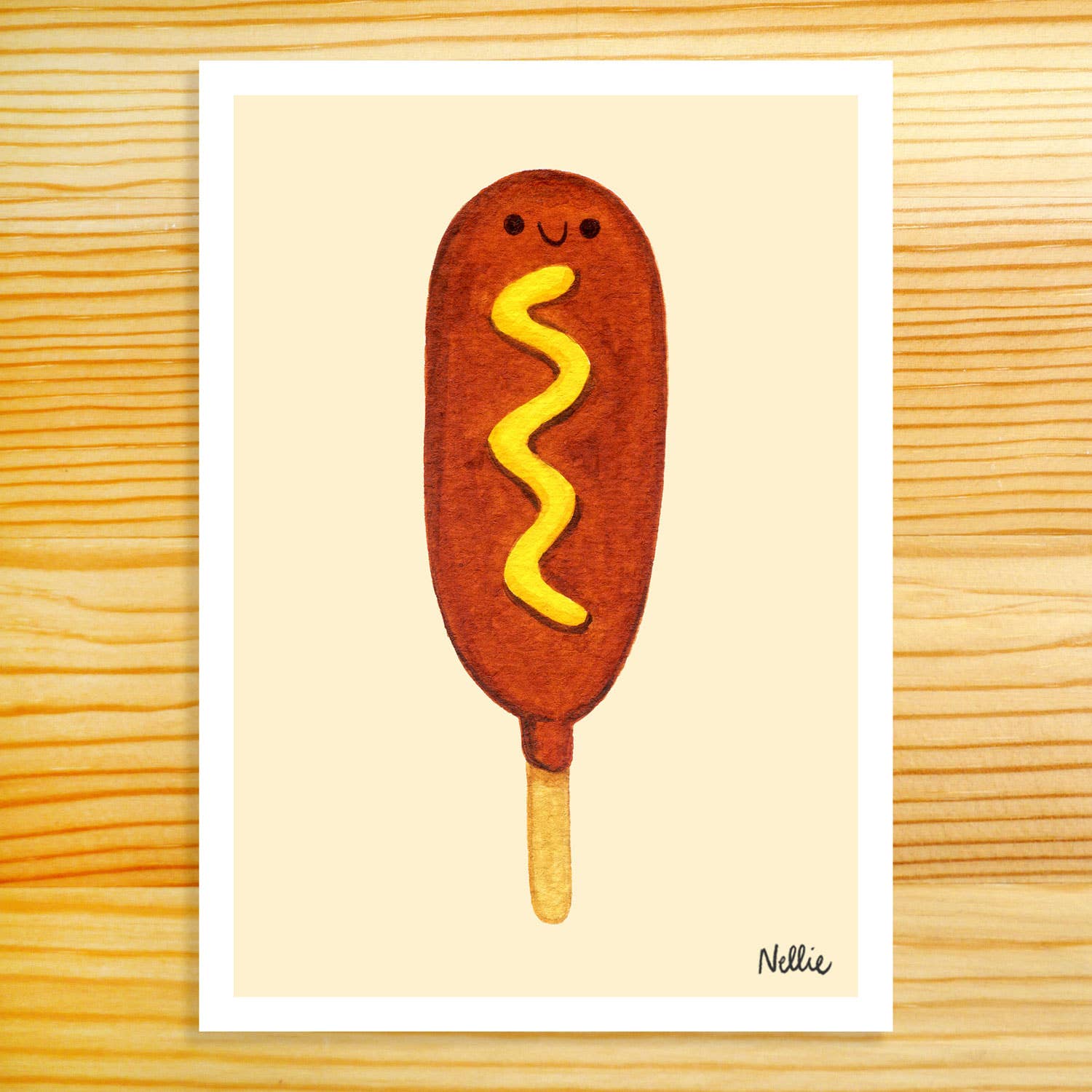 5x7in Art Print of a watercolor painting. Image depicts a corndog with a smiling face and a yellow squiggle of mustard. Background is light tan with a white border. Artist signature "Nellie" in the bottom right corner.