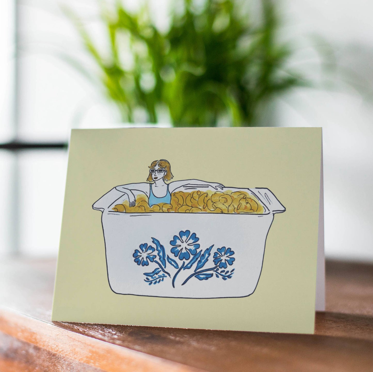 Greeting card with woman sitting in a mac and cheese filled vintage corningware dish
