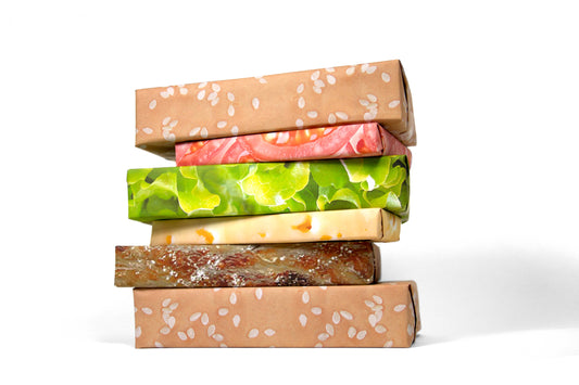 Stack of six boxes wrapped in cheeseburger wrapping paper, each box corresponding to a component of a cheeseburger. From top to bottom -- Bun wrapping paper, tomato wrapping paper, lettuce wrapping paper, cheese wrapping paper, patty wrapping paper and bun wrapping paper. 