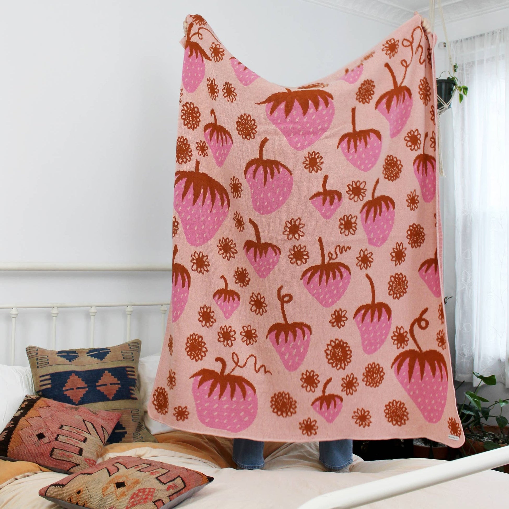 Knit blanket held up by a person on their bed --- majority of the blanket is light pink with different sized strawberries in a darker shade of pink and orange, along with flowers that are also orange all over it 