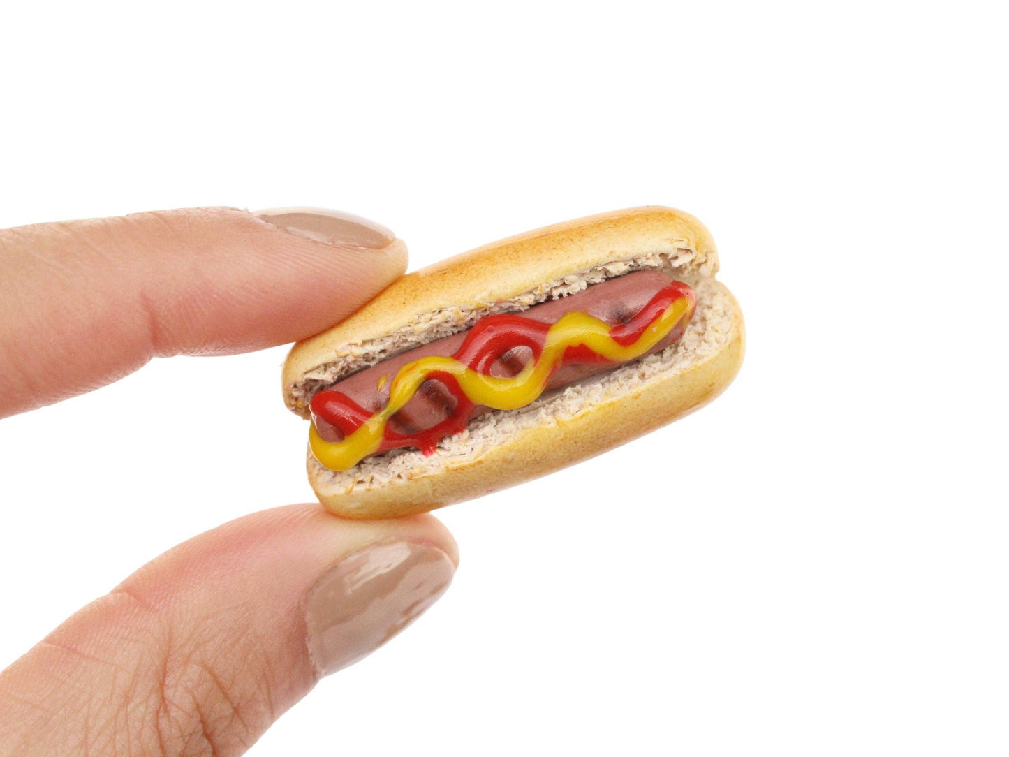 Photo of single mini hot dog magnet pinched between fingertips. Hot dog depicted with grill marks and a squiggle each of ketchup and mustard. White background.