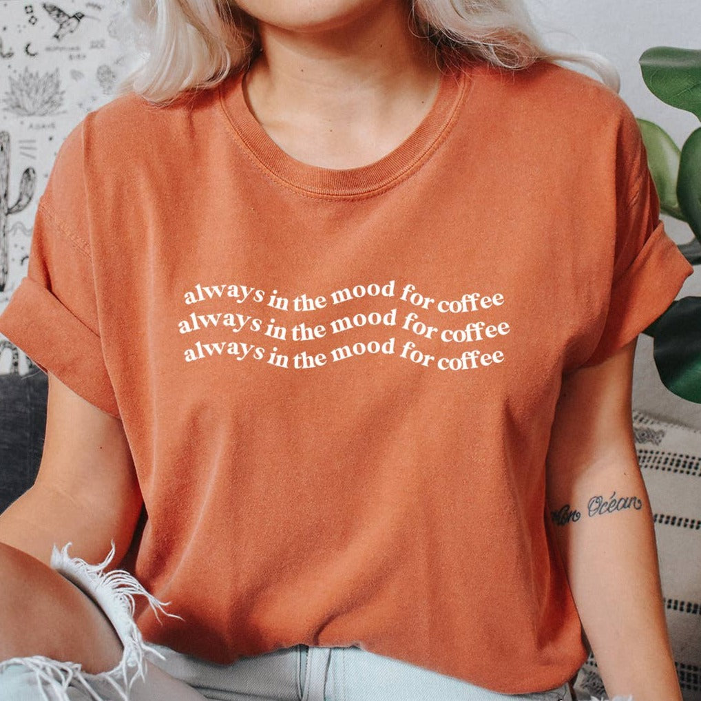 Vintage style tee-shirt in terracotta color, with wavy lines of white text "always in the mood for coffee" repeated three times.