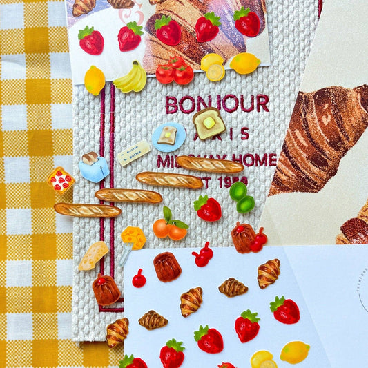 Sticker sheet with various fruits and types of bread on it -- includes but not limited to: strawberries, lemons, bananas, toast, butter, baguettes, croissants and caneles. 