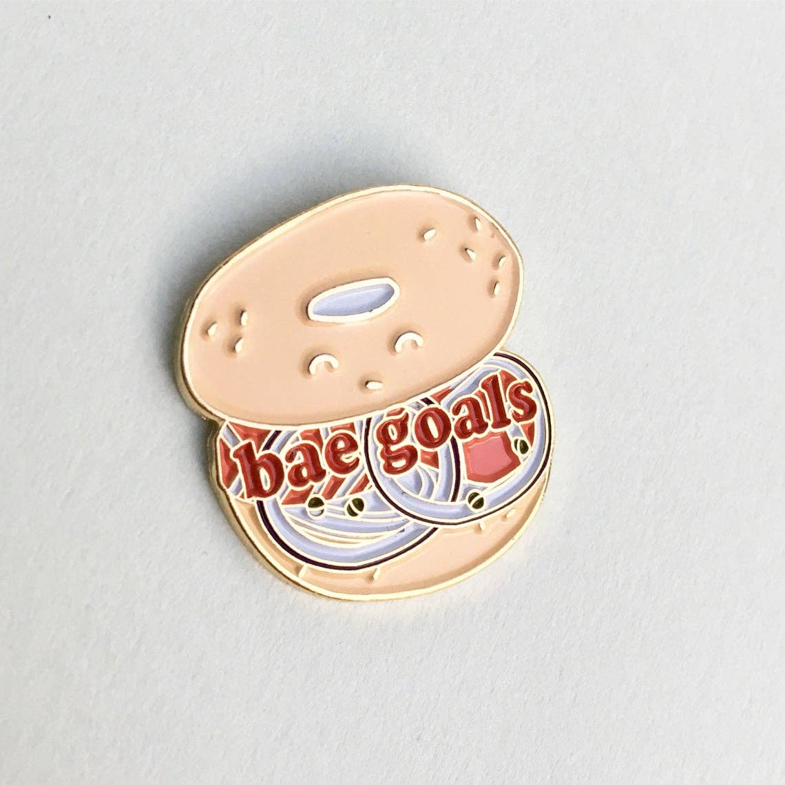 Bagel enamel pin that reads "Bae goals" on an open-faced bagel sandwich. 