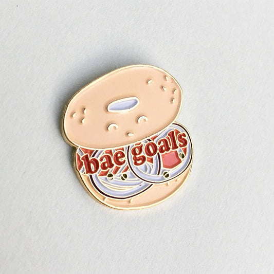Bagel enamel pin that reads "Bae goals" on an open-faced bagel sandwich. 