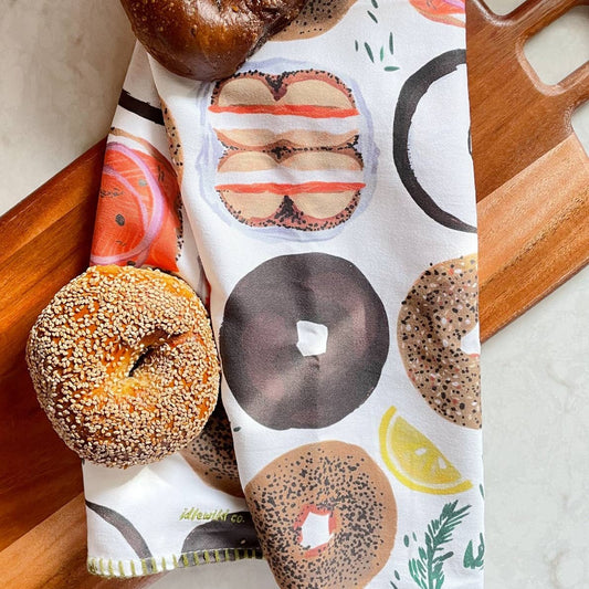 Tea towel with different types of bagels on it. It is laid out on a cutting board next to a sesame bagel and a pumpernickel bagel.  