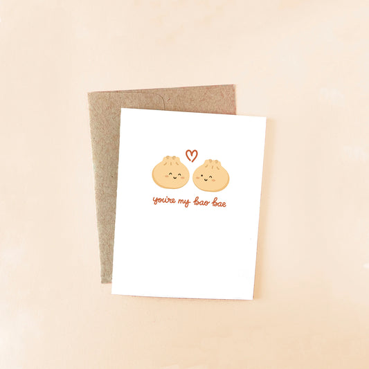 Greeting card with two bao buns next to each other and a red heart in between them, below them reads "you're my bao bae" in red cursive. All on white cardstock with a brown envelope
