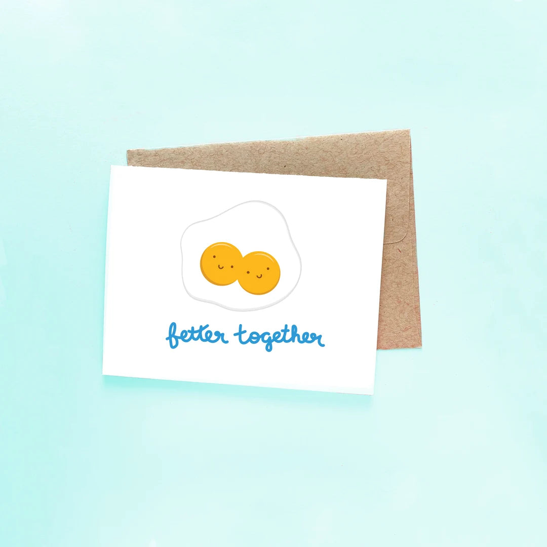 White greeting card that has an image of sunny side up eggs with two yokes, each with a smiley face and below it reads "better together" in blue cursive. Comes with a brown envelope. 