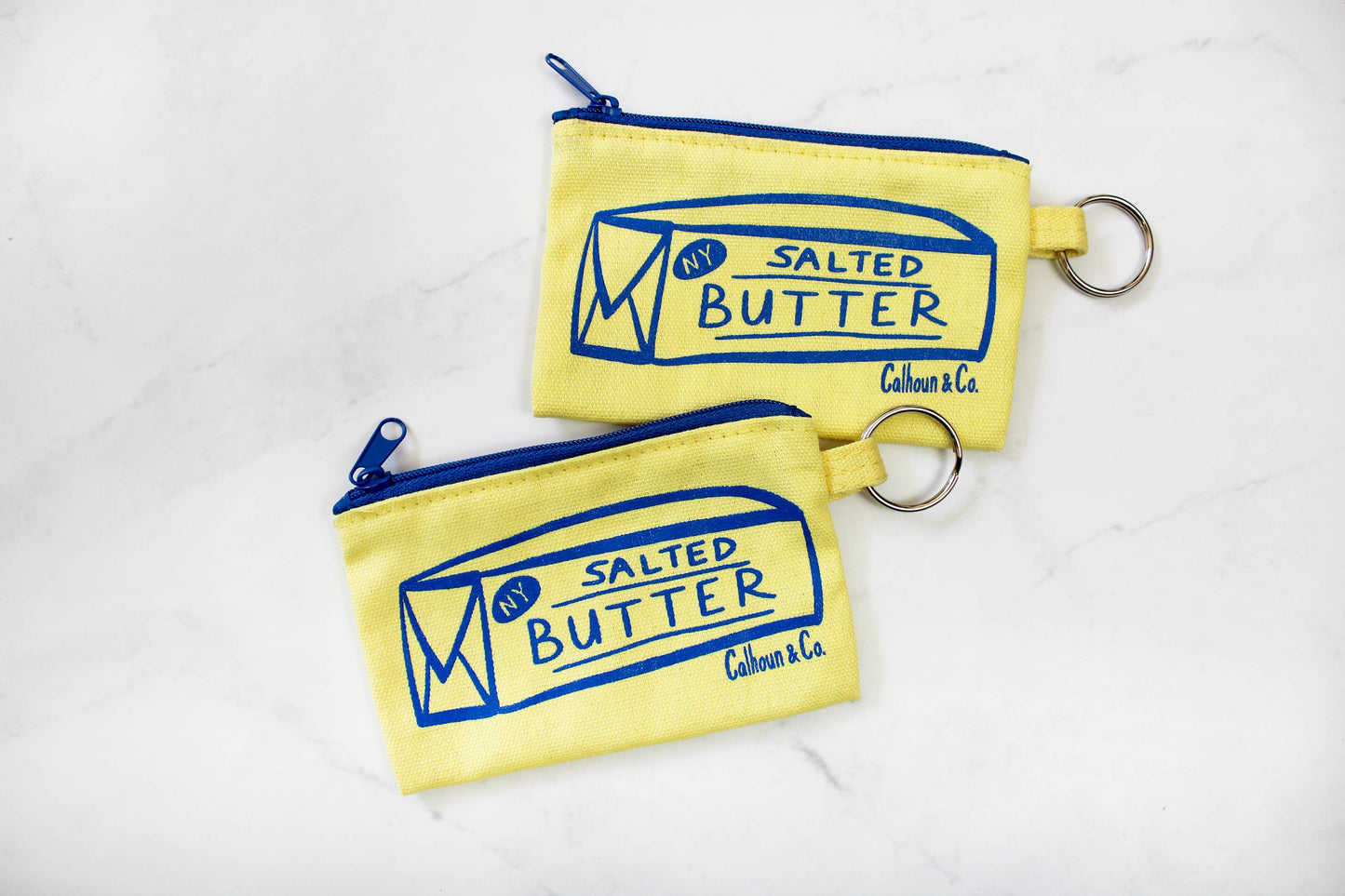 Two salted butter zippered pouches -- printed in blue ink on front is image of a stick of butter that reads "NY" in top left corner and "Salted Butter" in center 
