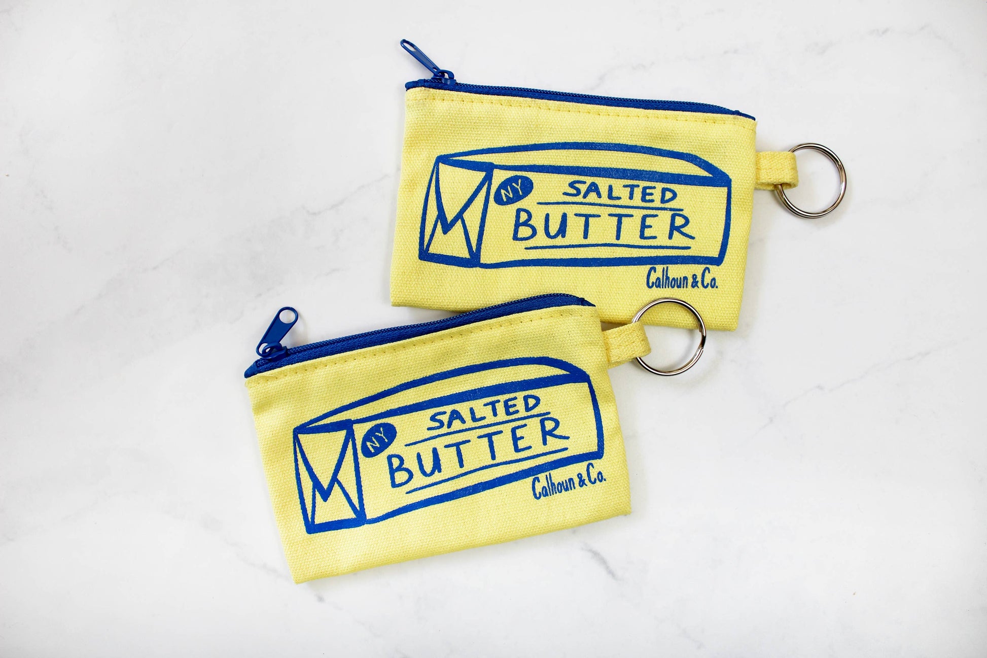 Two salted butter zippered pouches -- printed in blue ink on front is image of a stick of butter that reads "NY" in top left corner and "Salted Butter" in center 