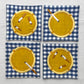 Set of 4 cotton coasters -- all are blue gingham with an embroidered yellow/gold ashtray on it. Each ashtray also has embroidered, variously smoked cigarettes on it. 
