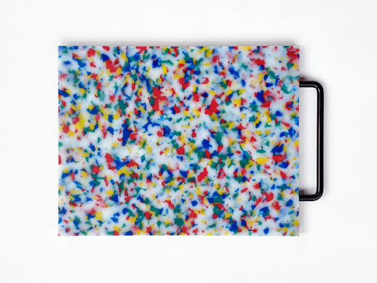 Cutting board in multi confetti -- red, yellow, blue and green confetti throughout the board. Handle is black. 