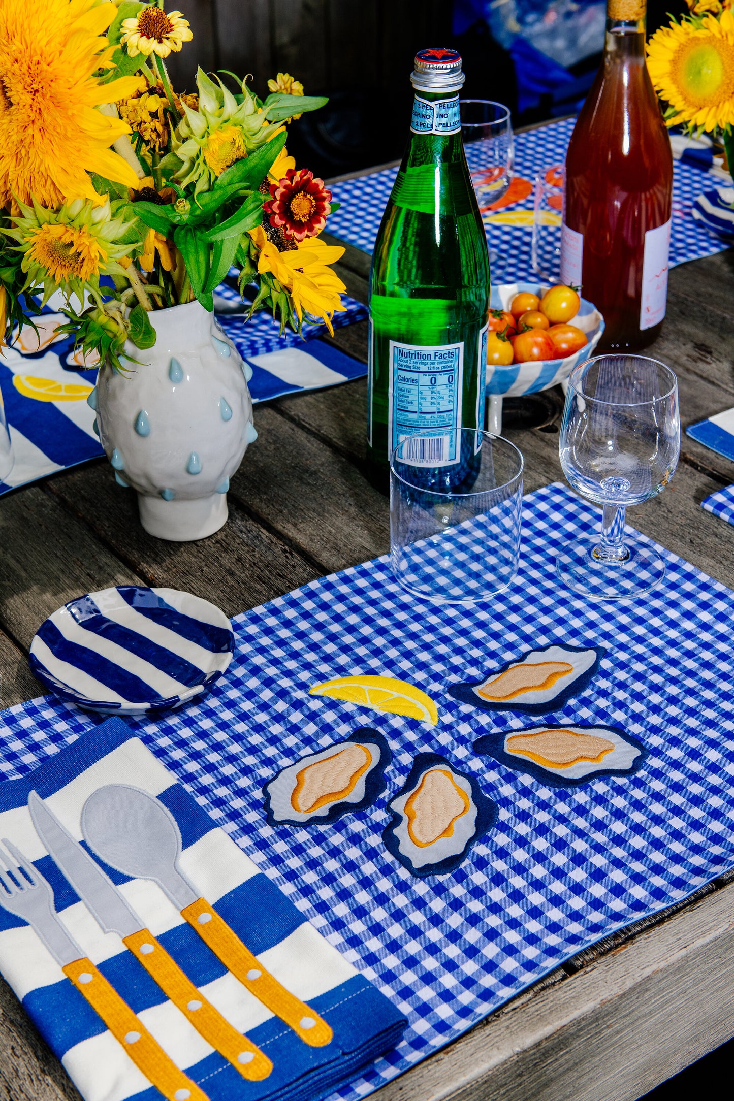 Table setting featuring a blue gingham placemat with four oysters on the half shell and a lemon wedge embroidered in the center 