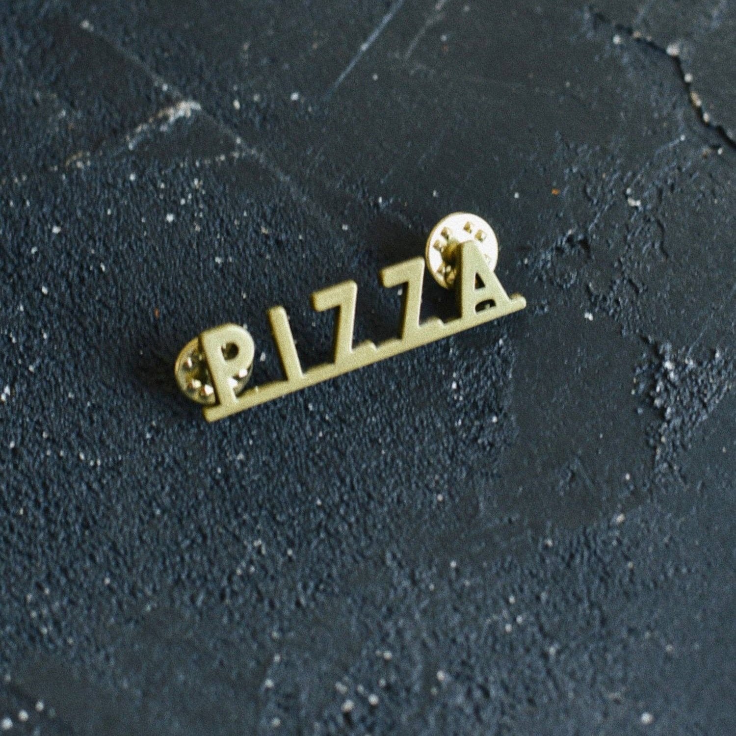 Brass colored enamel pin that spells out "pizza"