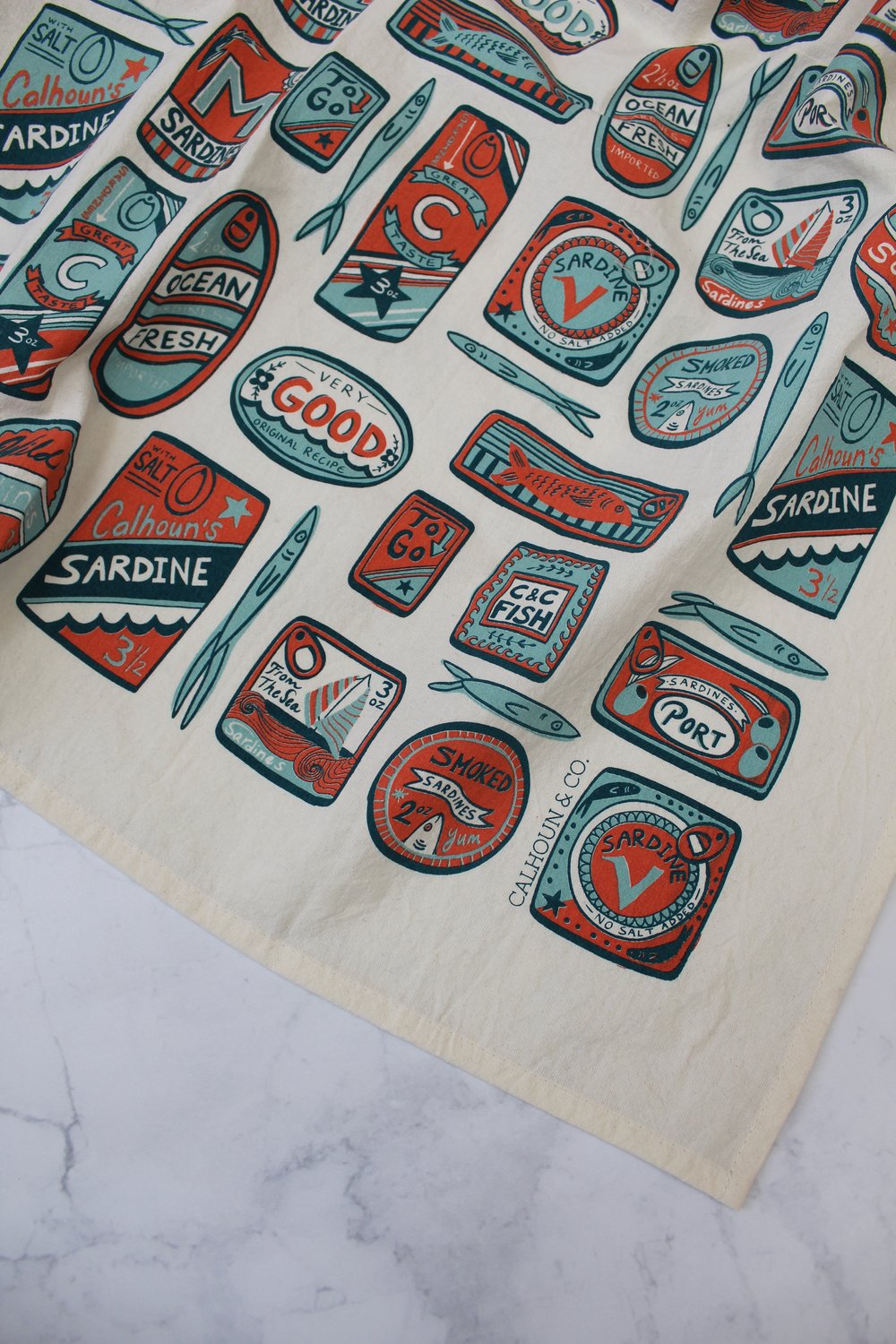 Close up view --- white tea towel with various shaped sardine tin cans in a red, light blue and navy colorway 