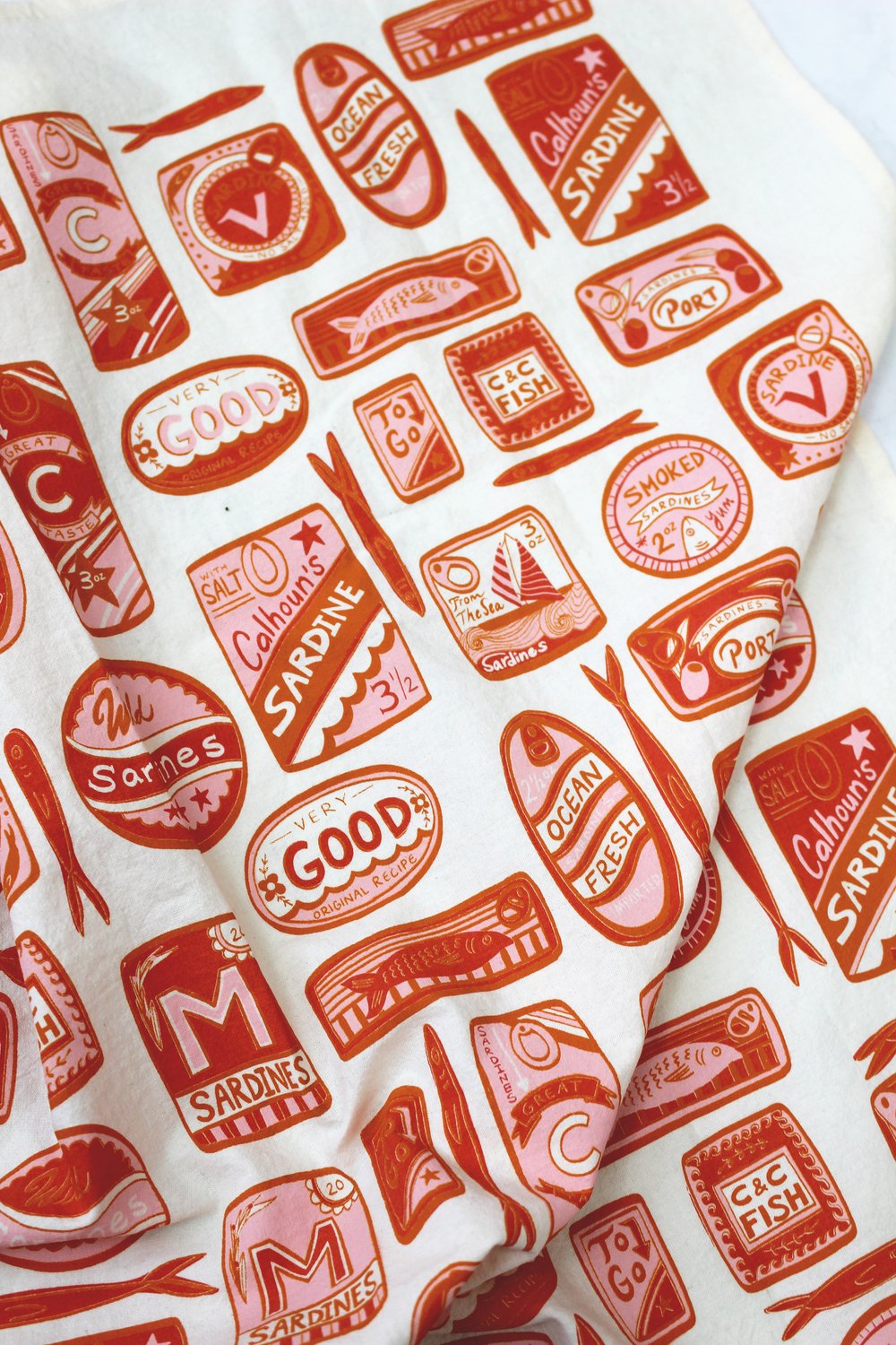 Close up view -- white tea towel with variously shaped sardine tin cans that are red and pink 
