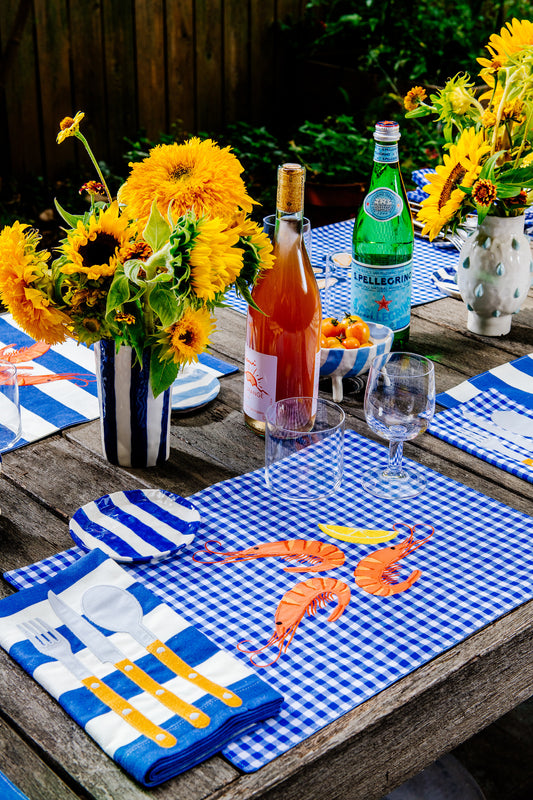Table setting featuring a blue gingham placemat with three shrimp and a lemon wedge embroidered in the center 