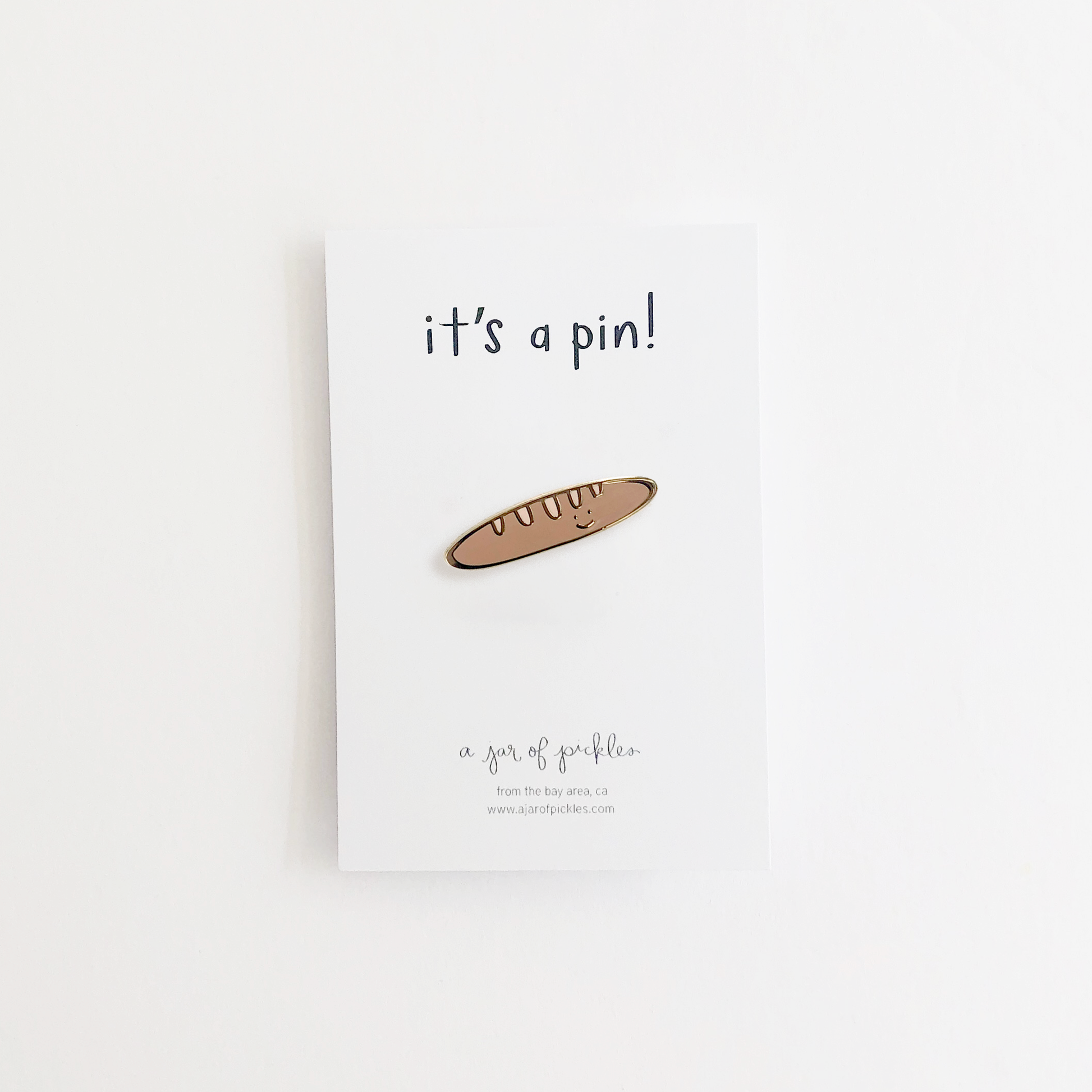 Enamel pin of a baguette with a smiley face, on top of a white card backing that says "it's a pin!"