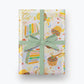 Birthday Cake Wrapping Paper shown with a blue bow. Illustration on wrapping paper features cupcakes and slices of colorful layer cake with lots of sprinkles. Multiple areas of text read "Make a WIsh!" and "Wishing you a very Happy Birthday" in gold script.
